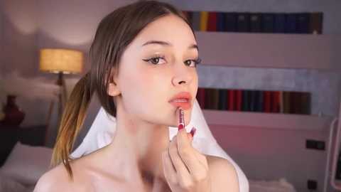 Video of a fair-skinned young woman with brown hair in pigtails, applying lipstick in a modern bedroom with colorful bookshelf and lamp.