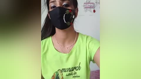 Media: Video of a young woman with medium brown skin, wearing a black face mask, green t-shirt with \"Mongarsh\" design, and pearl necklace. Background features a white wall and a red sign.