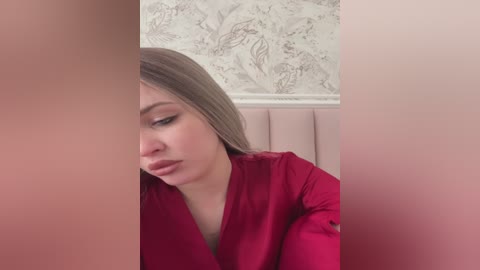 Media: Video of a fair-skinned woman with straight, light brown hair, wearing a red robe, seated in a room with a pink tufted headboard and a marble-patterned wallpaper.