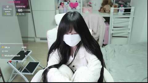 Media: Video of an Asian woman with long black hair and a white mask, sitting in a white chair in a clean, white-walled room, surrounded by laundry.