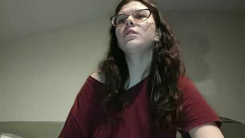 A video of a woman with long, curly brown hair, wearing glasses and a maroon off-shoulder top, standing indoors against a plain, off-white wall.