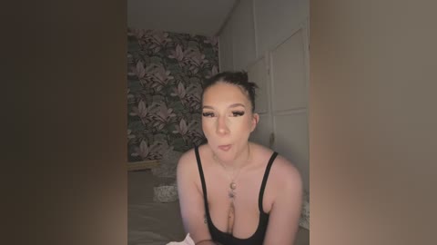Video of an East Asian woman with dark hair, fair skin, and large breasts, wearing a black tank top, blowing a kiss in a dimly lit bedroom with floral wallpaper.
