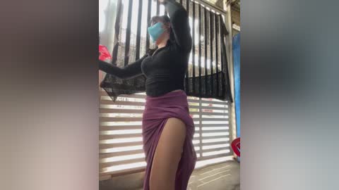 Video of a woman in a black top and purple skirt with a high slit, dancing on a balcony with a blue face mask, surrounded by metal bars and sunlight.
