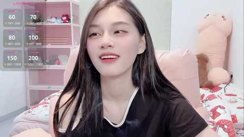 Video of a smiling young Asian woman with long black hair, wearing a black top, in a pink bedroom with a stuffed bear and shelves.