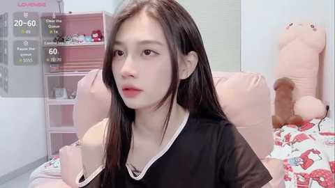 A video of an East Asian woman with long black hair, wearing a black top, sitting on a bed with pink pillows, in a room with a stuffed animal and pink decor.