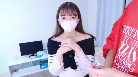 Video of an Asian woman in a black and white striped top, wearing a face mask, sitting on a couch in a modern living room with a computer monitor and a red-shirted man in the background.