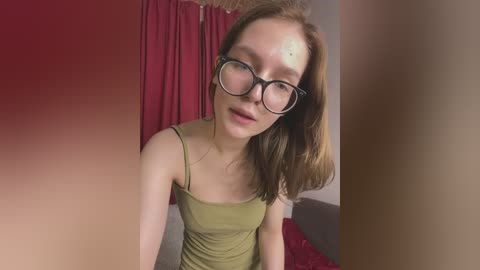 Video of a young, fair-skinned woman with shoulder-length auburn hair, wearing glasses, olive-green tank top, and red curtain backdrop, sitting on a bed.