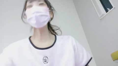 Video of a young Asian woman in a white t-shirt with black trim, wearing a surgical mask, standing in a sterile, white room.