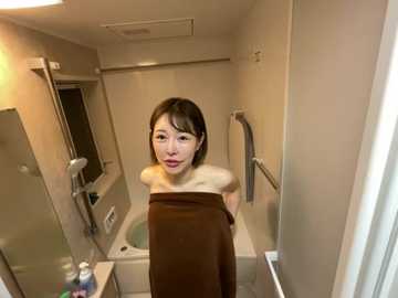 Video of a young Asian woman with fair skin and short black hair, wrapped in a brown towel, standing in a beige-tiled bathroom with a bathtub and shower.