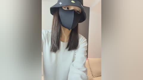 Video of a person wearing a black face mask, black bucket hat, and light grey long-sleeved shirt, standing in a room with beige walls and a beige couch, taken from a slightly angled perspective.