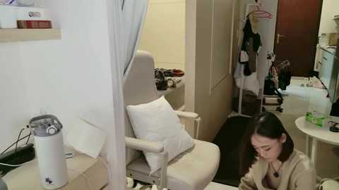 Video of a small, cluttered room with a beige armchair, white pillow, and a woman with long dark hair, wearing a beige coat, sitting with her hands over her face, surrounded by clothing and personal items.
