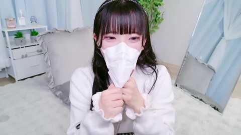 Video of an Asian woman with long black hair, wearing a white fur-trimmed robe and a white face mask, standing indoors with a white bed, green plant, and a mirror in the background.