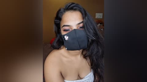 Video of a young woman with long black hair, wearing a black face mask, light grey off-shoulder top, and brown wall background.