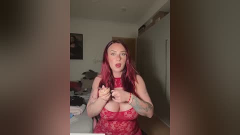 Video of a curvy, fair-skinned woman with red hair and tattoos, wearing a red lace lingerie top, posing in a dimly lit bedroom.