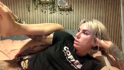 Video of a tattooed, platinum blonde woman with piercings, wearing a black t-shirt, lying on a bed, stretching her legs. Background shows a striped wall and a green plant.