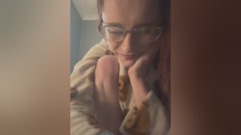 A video of a Caucasian woman with fair skin, glasses, and brown hair, sitting in a dimly lit room, leaning on her hand, wearing a beige sweater.