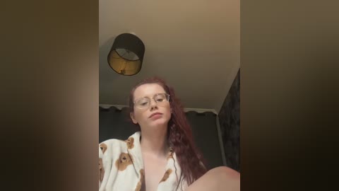 Video of a fair-skinned woman with long red hair, wearing glasses, a white robe with brown paw prints, and a relaxed expression, standing in a dimly lit room with a black lampshade hanging from the ceiling.