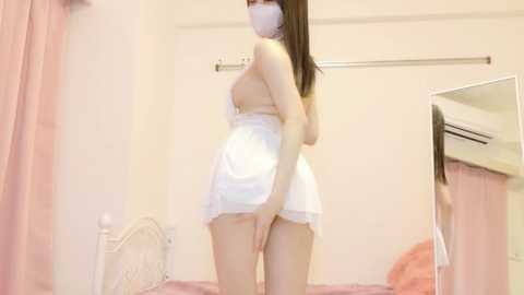 A video of an Asian woman with long, straight brown hair, wearing a white, lace-trimmed dress that reveals her buttocks, standing in a pink bedroom with a white bed and a mirror.