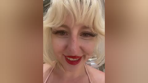 A video of a smiling woman with platinum blonde hair, red lipstick, and pale skin, wearing a light pink top, set against a blurred background.