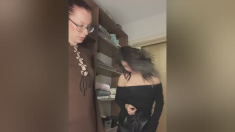 Video of a young woman with dark hair, wearing a black off-shoulder top, adjusting her top in a cluttered room.