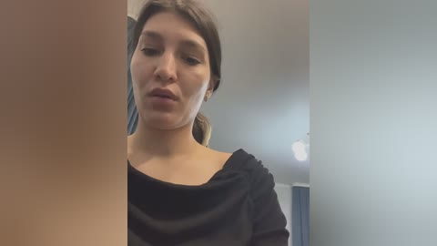 Video of a young woman with light skin and brown hair in a ponytail, wearing a black top, looking confused or in thought. Background shows a white ceiling and grey wall.