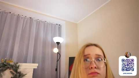 A video of a blonde woman with glasses, standing in a warmly lit room with beige walls, a white fireplace, and a grey curtain. A QR code in the bottom right corner indicates an Instagram profile.