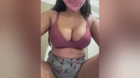 A video of a woman with medium skin tone, long dark hair, wearing a pink bra and high-waisted floral-patterned shorts. Her breasts are prominent, and she is partially obscured by blurred, pinkish hands at the edges of the image.