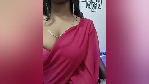 Video of a South Asian woman with medium brown skin, wearing a revealing, off-shoulder red sari, with a gold necklace. Background includes a white wall with black and white framed art and a partially visible bed.