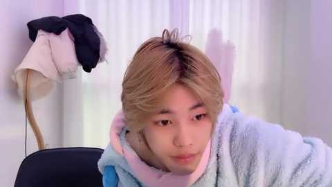 Video of a young Asian man with light brown hair, wearing a blue and pink plush robe, leaning forward, in a room with white curtains and a black chair.