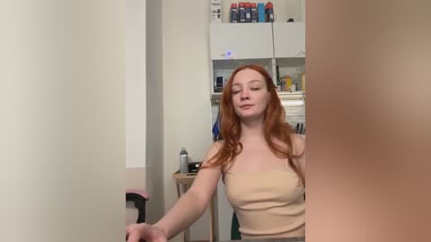 Video of a fair-skinned, red-haired woman with long wavy hair, wearing a beige strapless top, standing in a small, cluttered kitchen with white cabinets and a refrigerator.