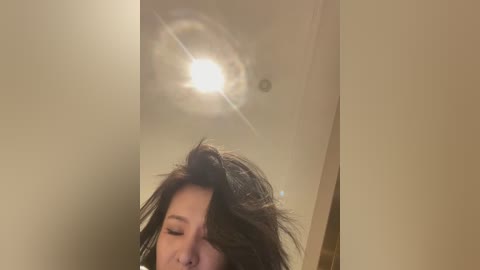 A video of an Asian woman with long, dark hair, partially covering her face, captured from the waist up. She appears to be in a dimly lit room with a ceiling light shining brightly in the background.