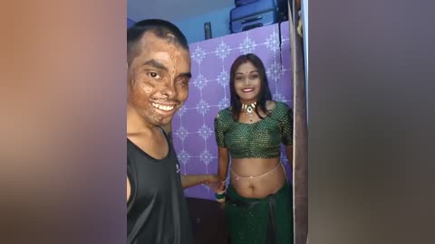 Video of a young South Asian man with a broad smile, standing next to a woman in a green lehenga choli. Background includes a pink patterned wall and a black doorway.