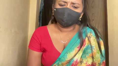Video of a South Asian woman with medium skin tone, wearing a black mask, red short-sleeved top, and colorful sari, standing in a doorway, eyes downcast.