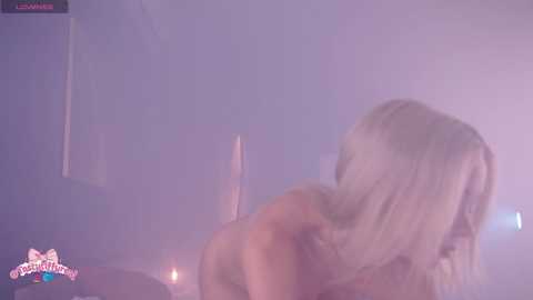 A foggy, dimly-lit video of a nude, light-skinned woman with platinum blonde hair, seen from behind, in a bedroom. The image has a soft, dreamy quality with a watermark in the bottom left corner.
