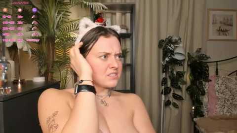 Video of a topless woman with a white cat headband, wearing a black wristwatch, in a cozy room with plants, a shelf, and a pink curtain.