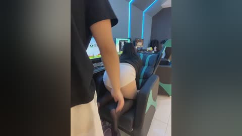 Video of a man, in black clothes, adjusting a woman's pants, revealing her buttocks, in a modern gaming room with blue lighting and multiple screens.