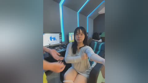 Video of an Asian woman with long black hair, wearing a light grey top, seated in a gaming chair, playing a video game, surrounded by computer screens and blue neon lights.