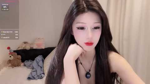 Video of a pale, long-haired East Asian woman with red lipstick, in a bedroom with stuffed animals and a bed.