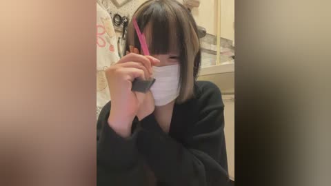 Video of an East Asian woman with short, straight black hair, wearing a black mask, and a black hoodie, using a pink hairbrush in a bathroom with beige tiles and a sink.