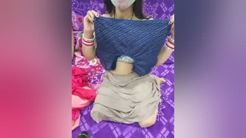 Media: Video of an Asian woman with light skin, kneeling on a purple bedspread. She wears a blue quilted jacket over a white bra, beige skirt, and red wristbands. Clothes are strewn around her.
