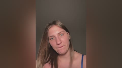 Video of a young Caucasian woman with long, straight brown hair, wearing a blue tank top, against a dark, blurred background. She has a neutral expression.
