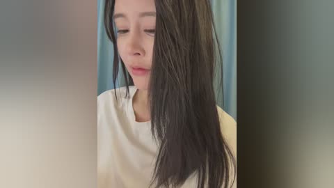 Video of a young Asian woman with long, wet, black hair, wearing a white t-shirt, standing in front of a blurred, muted blue background.