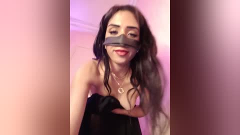 Video of a Latina woman with long dark hair, wearing a black strapless dress, blindfolded with a gray cloth, smiling seductively, in a dimly lit room with pink lighting.