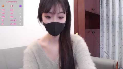 A video of an East Asian woman with long black hair, wearing a black face mask and a grey fuzzy sweater, sitting indoors with a calendar and floral curtains in the background.