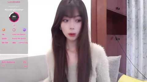 A video of a young Asian woman with long, straight black hair, wearing a beige fur coat, in a modern living room with a wooden cabinet and gray sofa.