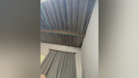 Video of a leaky roof with a green pipe running through it. The roof is made of corrugated metal, and a gray curtain hangs below. The room has white walls and a light fixture is visible.