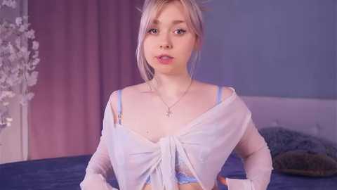 Video of a young, fair-skinned woman with platinum blonde hair, wearing a sheer white top and light blue bralette, indoors against a pink and blue backdrop.
