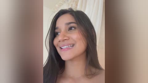 Video of a young woman with long, dark hair, light skin, and a warm smile. She is topless, standing against a beige curtain background.