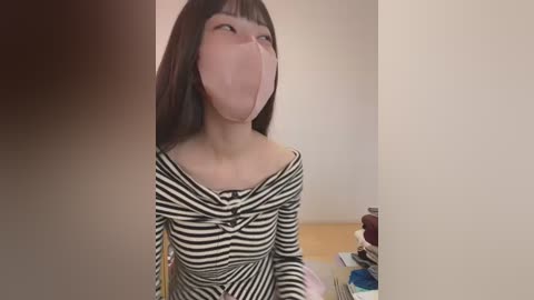 A video of a young woman with straight black hair and a mask, wearing a black and white striped off-shoulder top, sitting indoors with minimal background.