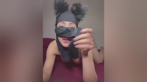 Media: Video of a woman with fair skin, wearing a black bear mask with fluffy ears, lying on a red bed, holding a black blindfold.